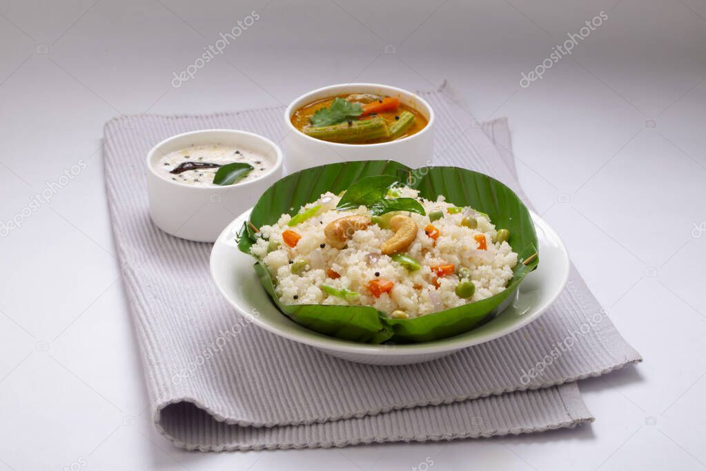 Upma made of samolina or rava , south indian breakfast item which is beautifully arranged in a bowl made of banana leaf and garnished with fried cashew nut on napkin with sambar and chutney as side dish on white textured background.
