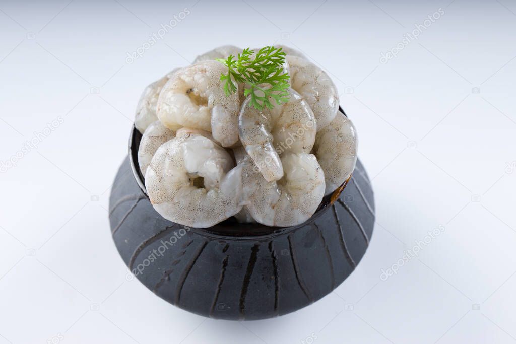 Raw peeled prawns or shrimp arranged in a black ceramic small pot and garnished with coriander leaf which is ready to cook ,placed on a white textured background.