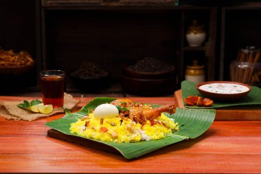 Kerala Tavuk dum biryani, geleneksel olarak muz yaprağı ve raita karışımı bir meze ve ahşap arkaplan veya dokuya sahip sıcak limon çayı, seçici bir odak noktası.