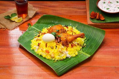 Kerala Tavuk dum biryani, geleneksel olarak muz yaprağı ve raita karışımı bir meze ve ahşap arkaplan veya dokuya sahip sıcak limon çayı, seçici bir odak noktası.