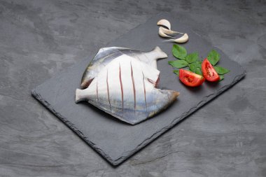 Raw white pomfret,ready to cook pomfret arranged on a rectangle shape graphite sheet with tomato ,curry leaves and garlic  as some cooking ingrediants on grey colour background. clipart