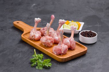 Çiğ tavuk lolipopu, altı parça tavuk lolipopu servis tahtasına yerleştirilmiş siyah biber, kişniş yaprağı ve arka planda gri desenli salatalık dilimleri.
