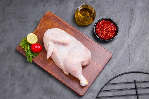 Raw half chicken with skin arranged on brown wooden board and garnished with parsely,small tomato,chilli flakes ,oil and lemon slices with delivery box on stone textured or graphite colour background,isolated