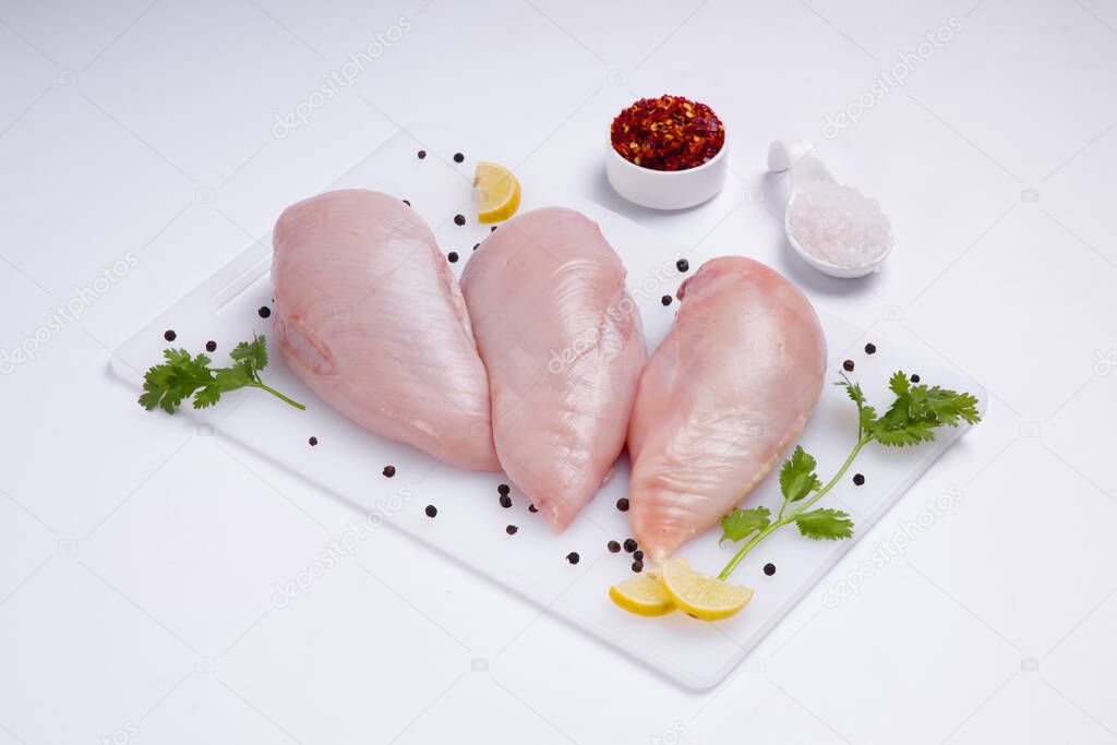 Raw chicken breast fillet without skin arranged on a white board and garnished with small lemon slices, a small bowl of chilli flakes and salt arranged on white coloured texture