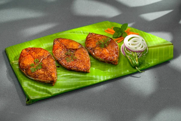 Seer fish fry arranged beautifully and garnished with onion, lemon and tomato slices on banana leaf covered base which is placed on solid green texture with abstract shadows on background.