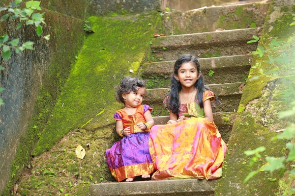 South Indian girl kids wearing  beautiful traditional dress long skirt and blouse,sitting on the long  steps of a house covered with moss green fungus, greenery background.