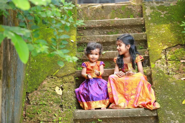 南インドの女の子の子供たちは 美しい伝統的なドレスの長いスカートとブラウスを着て 苔緑の真菌で覆われた家の長いステップに座って 緑の背景 — ストック写真