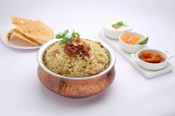 Veg Biriyani , garnished with fried onion ,coriander leaf and fried cashew nut in an antique brass vessel with lemon pickle ,veg salad, raitha and aplate of masala paapad as side dish with white background