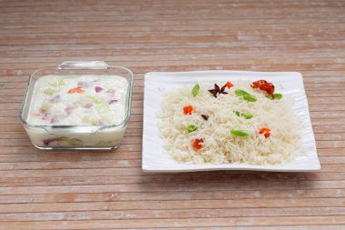 Basmathi pilavı, sebzeler ve Hint baharatları kullanılarak yapılan sebze pulav veya Pulao. Raita, ahşap arkaplan veya desen içeren kare beyaz bir sofra takımında yan yemek olarak düzenlenmiştir.