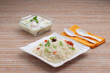 Basmathi pilavı, sebzeler ve Hint baharatları kullanılarak yapılan sebze pulav veya Pulao. Raita, ahşap arkaplan veya desen içeren kare beyaz bir sofra takımında yan yemek olarak düzenlenmiştir.