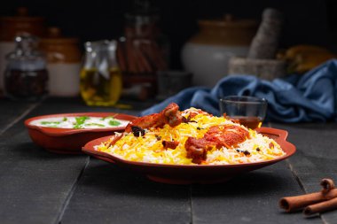 Mandi Biryani tavuğu, Tandır tavuğu, Hydrabadi tavuk biryani basmathi pilavı ve raita yanında siyah arkaplan ve desenli bir toprak tabaktaki sıcak limon çayı, seçici odak noktası.