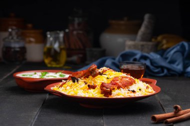 Mandi Biryani tavuğu, Tandır tavuğu, Hydrabadi tavuk biryani basmathi pilavı ve raita yanında siyah arkaplan ve desenli bir toprak tabaktaki sıcak limon çayı, seçici odak noktası.
