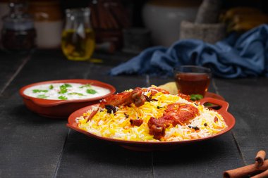 Mandi Biryani tavuğu, Tandır tavuğu, Hydrabadi tavuk biryani basmathi pilavı ve raita yanında siyah arkaplan ve desenli bir toprak tabaktaki sıcak limon çayı, seçici odak noktası.