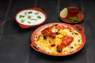 Mandi Biryani tavuğu, Tandır tavuğu, Hydrabadi tavuk biryani basmathi pilavı ve raita yanında siyah arkaplan ve desenli bir toprak tabaktaki sıcak limon çayı, seçici odak noktası.