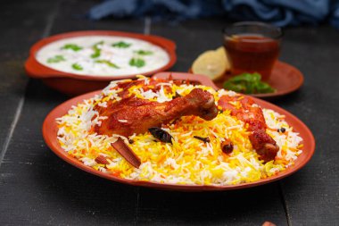 Mandi Biryani tavuğu, Tandır tavuğu, Hydrabadi tavuk biryani basmathi pilavı ve raita yanında siyah arkaplan ve desenli bir toprak tabaktaki sıcak limon çayı, seçici odak noktası.