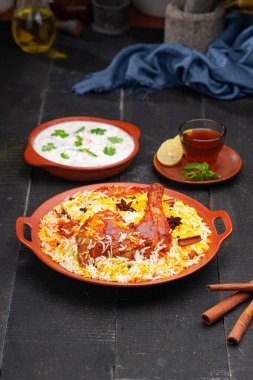 Mandi Biryani tavuğu, Tandır tavuğu, Hydrabadi tavuk biryani basmathi pilavı ve raita yanında siyah arkaplan ve desenli bir toprak tabaktaki sıcak limon çayı, seçici odak noktası.