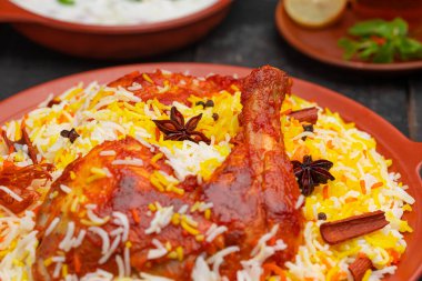 Mandi Biryani tavuğunun yakın plan görüntüsü, Basmathi pilavı ile Hydrabadi tavuk biryani ve raita yanında siyah arkaplan veya desenli bir toprak tabaktaki sıcak limon çayı, seçici odak noktası.