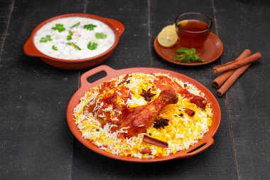 Mandi Biryani tavuğu, Tandır tavuğu, Hydrabadi tavuk biryani basmathi pilavı ve raita yanında siyah arkaplan ve desenli bir toprak tabaktaki sıcak limon çayı, seçici odak noktası.