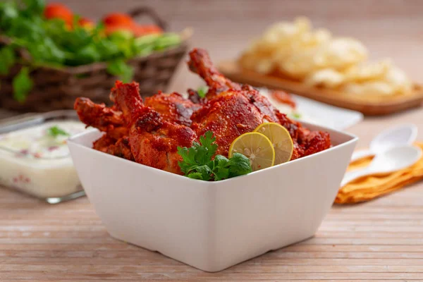 Spicy Tandoori chicken garnished with coriander leaf in square shaped white bowl with veg pulav, raita and paapad in background with wooden texture,selective focus