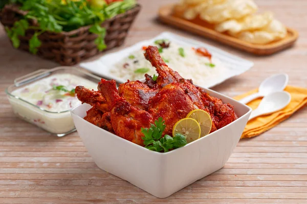 Spicy Tandoori chicken garnished with coriander leaf in square shaped white bowl with veg pulav, raita and paapad in background with wooden texture,selective focus