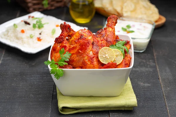 Tandoori chicken garnished with lemon and coriander leaf in square shaped white bowl with veg  pulav, raita and paapad in background with black texture,selective focus