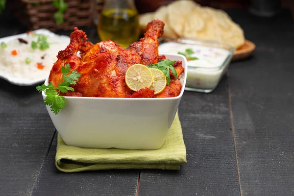 Tandoori chicken garnished with lemon and coriander leaf in square shaped white bowl with veg  pulav, raita and paapad in background with black texture,selective focus