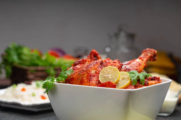 Tandoori chicken garnished with lemon and coriander leaf in square shaped white bowl with veg  pulav, raita and paapad in background with black texture,selective focus