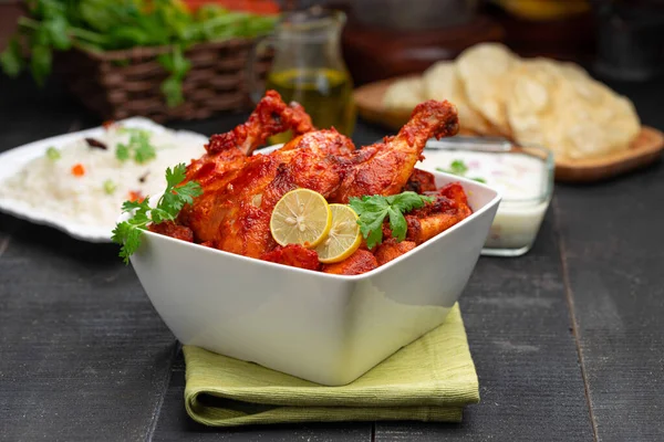 Tandoori chicken garnished with lemon and coriander leaf in square shaped white bowl with veg  pulav, raita and paapad in background with black texture,selective focus