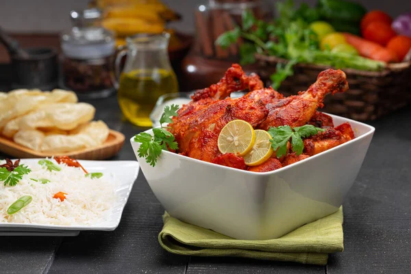 Tandoori chicken garnished with lemon and coriander leaf in square shaped white bowl with veg  pulav, raita and paapad in background with black texture,selective focus