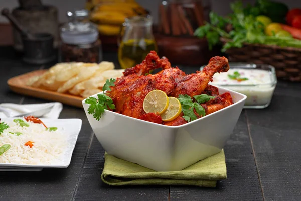 Tandoori chicken garnished with lemon and coriander leaf in square shaped white bowl with veg  pulav, raita and paapad in background with black texture,selective focus