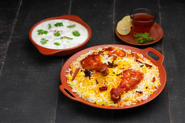 Mandi Biryani tavuğu, Tandır tavuğu, Hydrabadi tavuk biryani basmathi pilavı ve raita yanında siyah arkaplan ve desenli bir toprak tabaktaki sıcak limon çayı, seçici odak noktası.