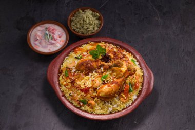 Jeera pilavı ve baharat kullanarak tavuk dhum biryani gri arka planda raitha ve limon turşusu eşliğinde toprak malzemesi..