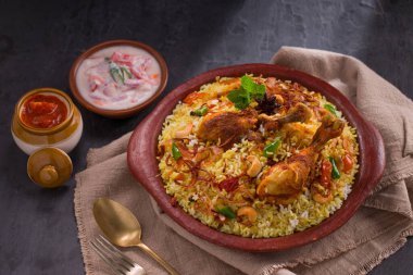 Jeera pilavı ve baharat kullanarak tavuk dhum biryani gri arka planda raitha ve limon turşusu eşliğinde toprak malzemesi..