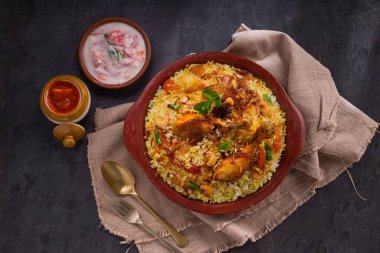 Jeera pilavı ve baharat kullanarak tavuk dhum biryani gri arka planda raitha ve limon turşusu eşliğinde toprak malzemesi..