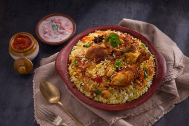 Jeera pilavı ve baharat kullanarak tavuk dhum biryani gri arka planda raitha ve limon turşusu eşliğinde toprak malzemesi..