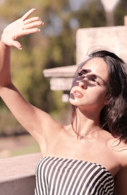 Gün batımında şehirde yürüyen koyu renk saçlı genç bir kadın. Güneş ışığından gözleri kamaşmış. Yüzünü koruyor.