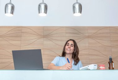 Diş hekimliği resepsiyonunda çalışan genç bir kadın not defterine notlar alır ve gülümser.