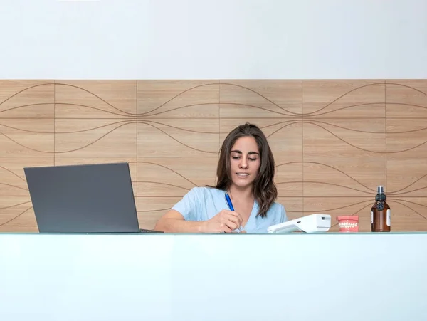 Diş hekimliği resepsiyonunda çalışan genç bir kadın not defterine notlar alır ve gülümser.