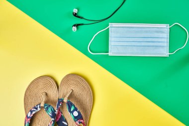 Stüdyo fotoğrafı, iki renk sarı ve yeşil dikey arka plan üzerinde köşegen yaz sandaletleri, kulaklıklar ve yüz maskesi tatil için hazır.