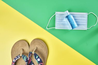 Stüdyo fotoğrafı, iki renk sarı ve yeşil dikey arka plan üzerinde köşegen yaz sandaletleri, antiseptik jel ve yüz maskesi tatil için hazır.