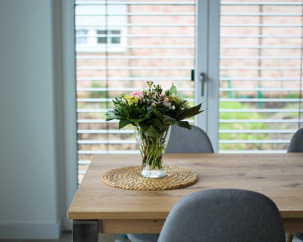 Свежие цветы в прозрачной стеклянной вазе на деревянном столе в гостиной. Минимализм домашней концепции. Белая стена и окно на фоне венецианских жалюзи.