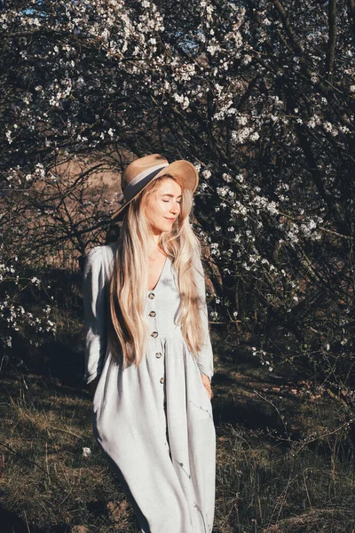 Blonde long hair caucasian white woman girl outdoor lifestyle portrait in spring romantic aroma nature park around bloom blooming trees and blossom tree sakura flowers pinky tones young in dress and hat