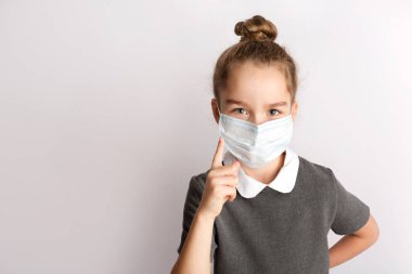 Katı bir okul elbisesi içinde çekici küçük bir kız, beyaz bir arka planda duruyor, elleriyle jestler yapıyor, yüzündeki mutlu ifadeye hayran.