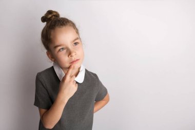 Katı bir okul elbisesi içinde çekici küçük bir kız, beyaz bir arka planda duruyor, elleriyle jestler yapıyor, yüzündeki mutlu ifadeye hayran.