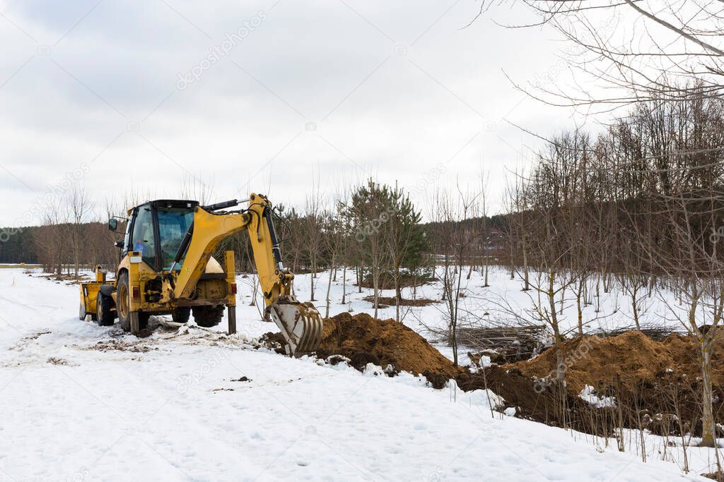 A yellow tractor excavator digs the ground for the repair and laying of underground utilities and cables for electricity and communications. During the winter season.