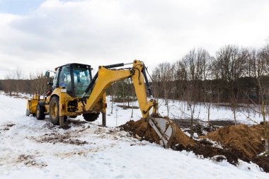 Sarı bir traktör kazıcısı, elektrik ve iletişim için yer altı tesisleri ve kablolarının onarımı ve döşenmesi için toprağı kazıyor. Kış mevsiminde.