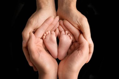 Anne ve baba, yeni doğan bebeğin çıplak ayaklarını siyah bir zeminde tutarlar. Bebeklerin küçük ayakları. Aile, aileler ve ev ödevi konsepti. Sağlık hizmetleri, pediatri.
