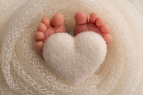 Ноги новонародженої дитини загорнуті в в'язану ковдру. Пальці новонародженої дитини тримають біле в'язане серце . — стокове фото