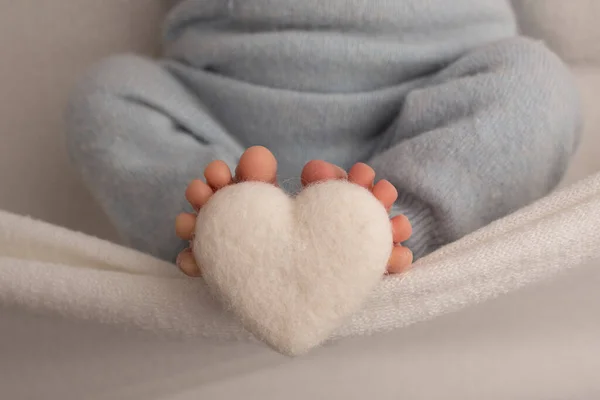 Ноги новонародженої дитини загорнуті в в'язану ковдру. Пальці новонародженої дитини тримають біле в'язане серце . — стокове фото