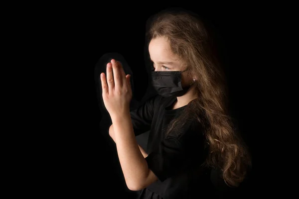 Nastoletnia dziewczyna w czarnej masce, chroniona przed koronawirusem, złożyła ręce w modlitwie. Czarny tło studio. — Zdjęcie stockowe